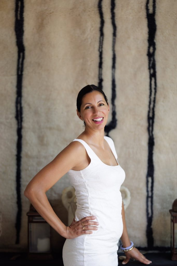 Nedjma Sahraoui souriante, main sur la hanche, en robe blanche devant une tapisserie berbère